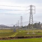 View of electric transmission towers stretching across a rural field with mountains in the background.