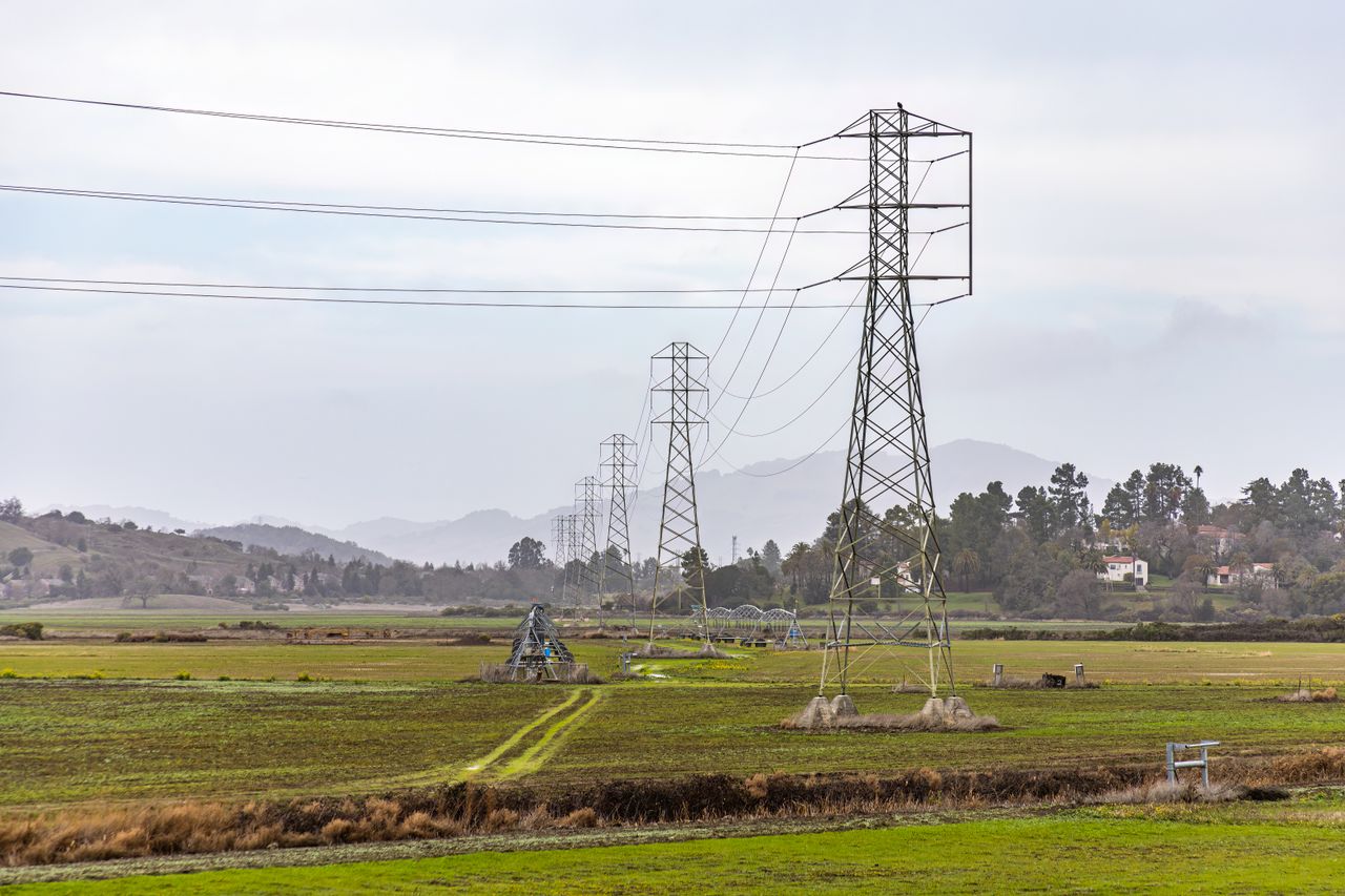 View of electric transmission towers stretching across a rural field with mountains in the background.
