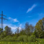 Power lines stretch across a vibrant rural landscape with a clear blue sky and green trees.